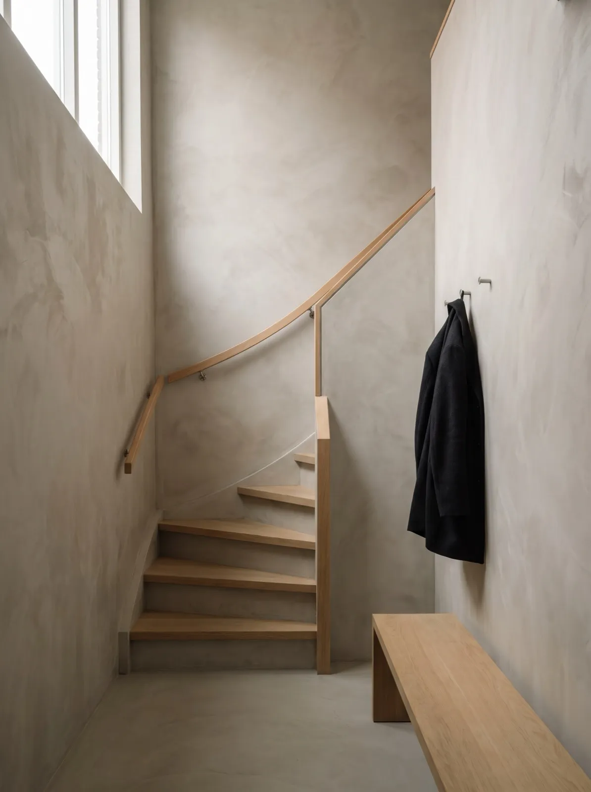 Hal en trapgat met wanden in microcement, mat en naadloos, met strijklicht van een hoog raam